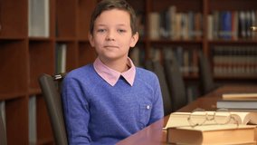 Footage portrait shot of the cute schoolboy sits near the bookshelf in the library. 4k video