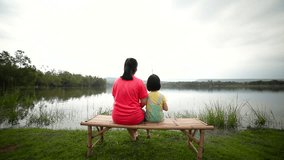 Asian mother with her daughter side by side on bamboo litter. Happy girl pulling rod in hands while fishing against landscape of lake. Concept of connecting children with nature. Travel on vacation.