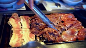 This close up video shows assorted meats sizzling and cooking on a korean bbq grill as they are moved around with tongs.