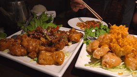This close up panning video shows a group of people gathering together over a feast of Chinese dishes as they server themselves with chopsticks.