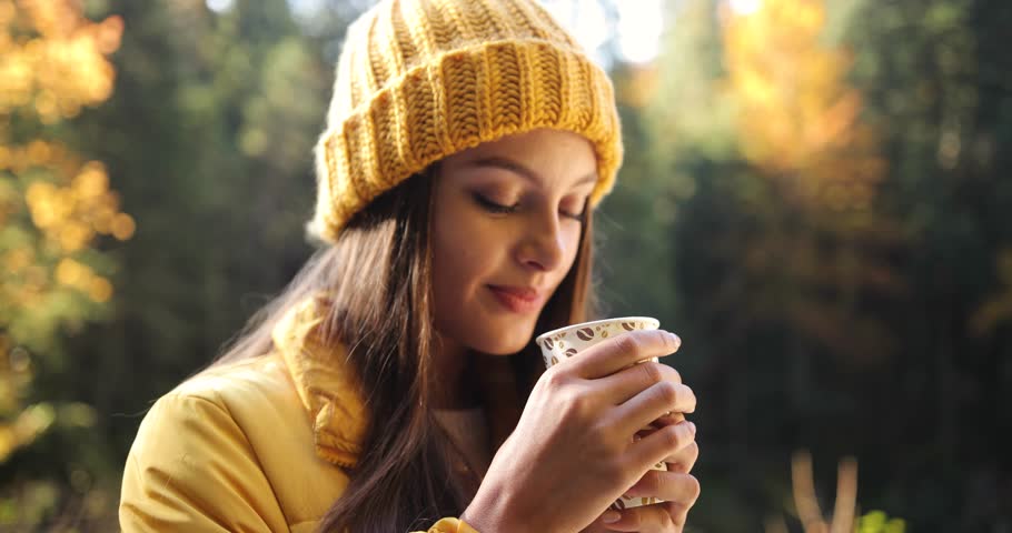 Female tourist in the mountains. Sun shines over stunning woman in yellow clothes while she enjoys hot coffee or tea in the forest
