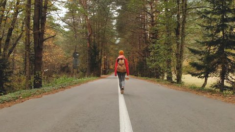 A young slim girl in a yellow hat and a vintage backpack walks along an asphalt road in the autumn yellow coniferous forest. Back view
