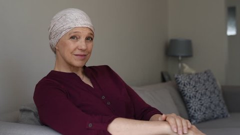 Mature woman with cancer in pink headscarf smiling sitting on couch at home and looking away. Mid woman suffering from cancer sitting after taking chemotherapy sessions looking at camera.