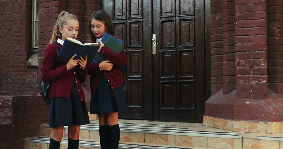Two Caucasian pretty teen girls standing outside the school with a book and reading it while children coming out from the school after lessons.