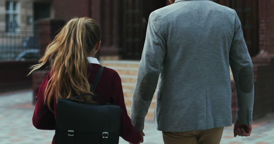 Back view on the caucasian father walking with his daughter hand in hand when bringing her to school in the morning. Rear.
