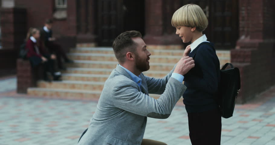 Portrait of the handsome father saying something to his son and hugging him in the morning in front of the school, then boy running to lessons and dad leaving for work. Royalty-Free Stock Footage #1018088053
