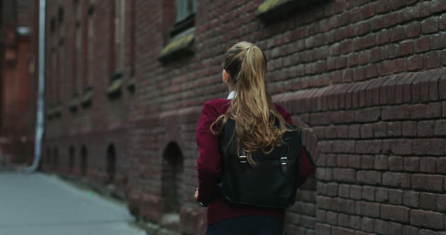Back view on the Caucasian schoolgirl in the uniform and with a bag walking the street after classes at school. Close up. Rear.