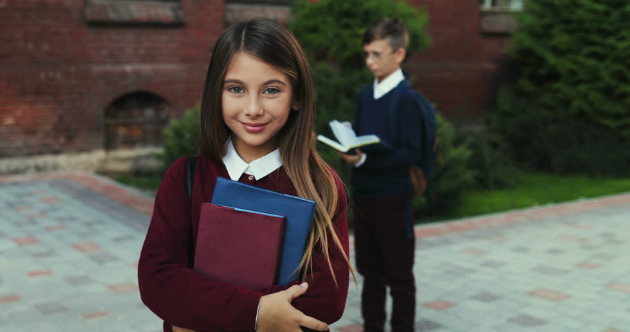 Camera zooming in the cheerful schoolgirl with books turning head to the camera and smilling while standing at the school. Other kids on the background.