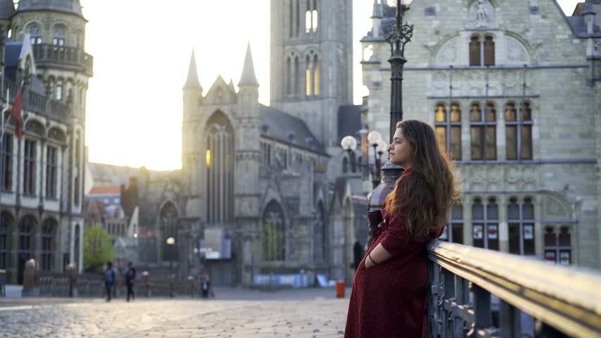 Young girl on St. Michael Bridge in sunny morning. Woman leaned against the handrails drinking coffee and go away, majestic cathedrals and towers at blurred background. Ghent, Belgium