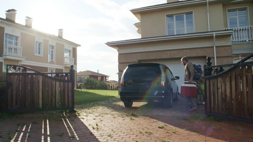 Panning wide shot of family preparing to go on picnic. Man and woman with basket and electric cooler and children with blanket and umbrella loading things into trunk of car parked in front of house Royalty-Free Stock Footage #1018105624