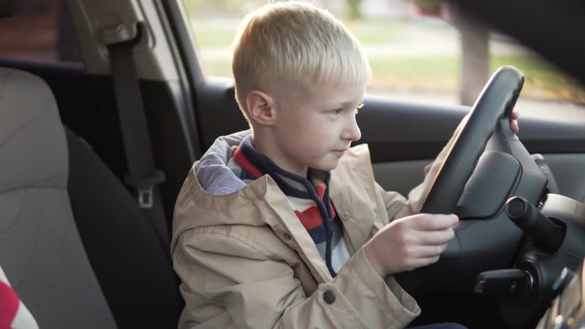 The boy sat in the driver's seat in the car. Drives behind the wheel and represents that he is a driver. The child is happy. He is trying to drive away. Closes the side windows. Royalty-Free Stock Footage #1018105654