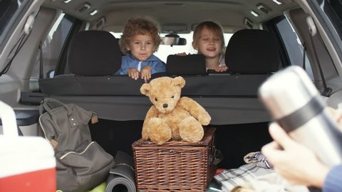 Medium shot of cute little boy with curly hair and cheerful girl sitting in backseat of family car and talking to parents putting picnic essentials in trunk before going on trip