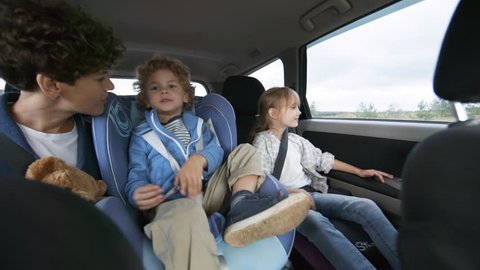 Handheld shot from inside of moving car: happy mother sitting in backseat giving plush toy to cute curly boy in child seat, then kissing him with affection. Curious little girl looking out window