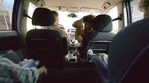 Shot from inside of moving car: happy young mother holding plush toy and playing with unrecognizable preschool-age boy in child seat and his elder sister riding in backseat on family road trip