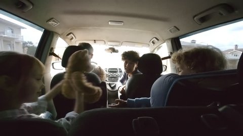Handheld shot from inside of moving car: smiling girl riding in backseat giving plush toy to little brother in child seat, then high-fiving happy mother in passenger seat