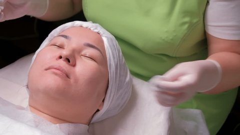 Beautician in a green dress wipes the face of an oriental girl with cotton pads. An asian muslim woman in a beauty salon is lying during a rejuvenating skin procedure. Women's hands in white gloves.