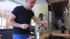 Woman with baby in arms take water bottle from refrigerator and man taste soup food in steamy pot. Happy family together in kitchen. Static shot. 4K UHD video clip.