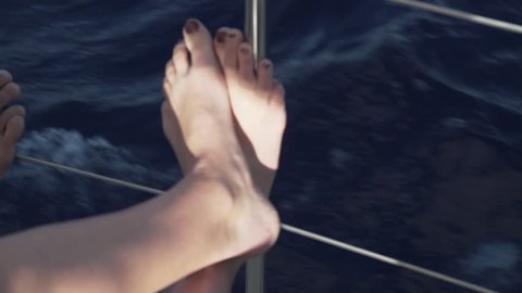 Feet resting on sailboat yacht railing while underway with blue water passing behind