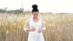 Young pretty pregnant woman posing on nature background. Expectant mother waiting for a baby. She holds and strokes her tummy with love. Girl have unusual hairstyle - braids