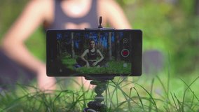 Young brunette woman vlogger shooting workout process video for her blog in a park with smartphone