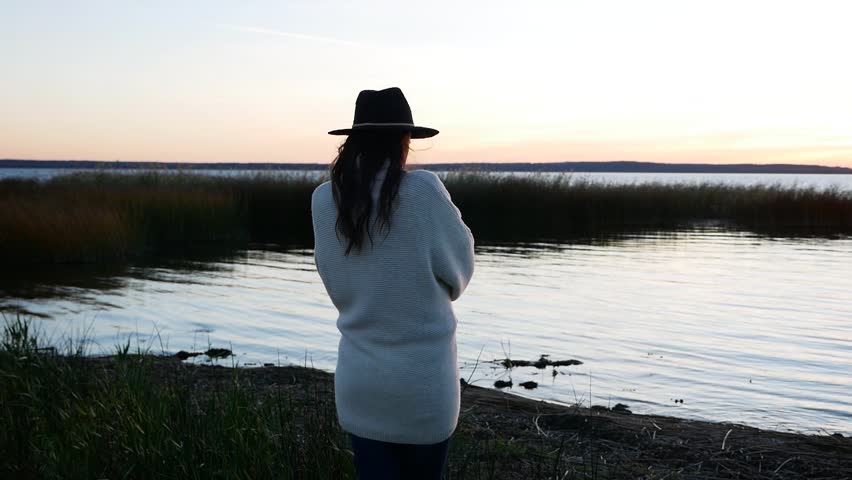 girl in a hat and a white sweater at sunset walking by the lake