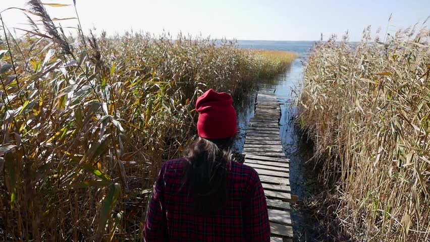 girl in a hat and sweater goes along the pier forward at the lake among the dry grass