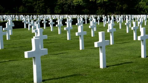 Pan-Grassy field of sun-lit of white crosses contrasting with dark trees, mark the graves of American dead at the WWII American Cemetery near Cambridge, England