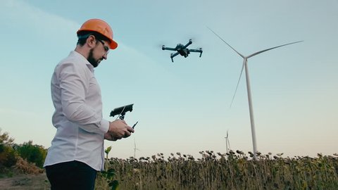 Caucasian Engineer in an orange protective helmet and goggles with a control panel controls the operation of a smart quadcopter drone for installing and building a wind power generator in a green