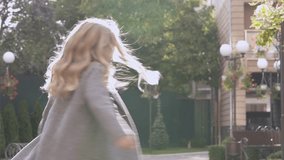 A pretty fair-haired girl in a grey coat twirls in the yard smiling and looking into the camera. The second girl runs towards her and the camera.