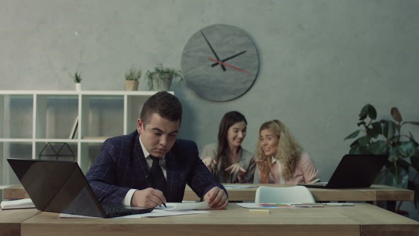Chatting giggling female office worker whispering gossips about male colleague in office. Two businesswomen bullying sad coworker and gossiping behind his back at workplace. Royalty-Free Stock Footage #1018149565