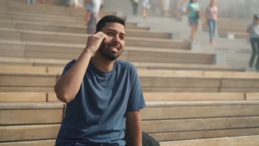 Bearded young man wearing blue t shirt sitting on stairs outside and talking on the phone. Slider slow motion medium shot