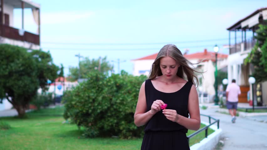 Curious busy young blonde female in black casual clothes looking closely at colorful pink tropical flower in hands