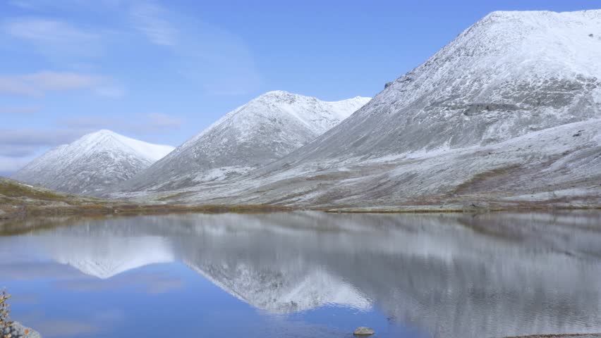backside view man in bright blue warm jacket watches fantastic highland lake reflecting snowy mountains and endless sky