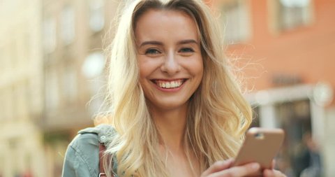 Portrait of the attractive blonde young woman chatting on her smartphone while being in the town street and then lookingto the camera. Close up. Outside.