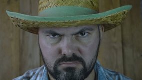 Adult unshaven man. Frown. Scary view. Close-up portrait. Straw hat. Wooden background. 4K video.