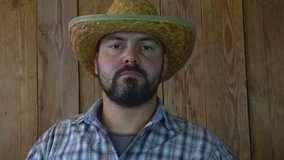 Adult unshaven man. Kind of boring. Looks straight. Straight face. Not to smile. Straw hat. Wooden background. 4K video.