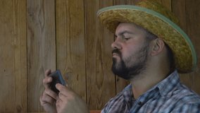 Adult unshaven man. Looks at the phone. Pensive look. Busy with work. Straw hat. Wooden background. 4K video.