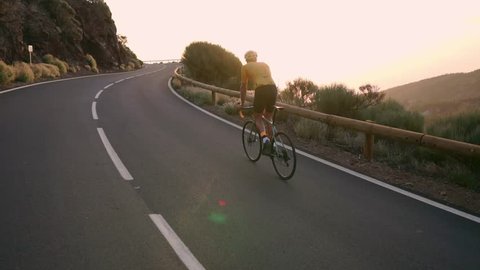 A professional cyclist in a helmet and sports equipment rides on a mountain highway at sunset in slow motion. Steadicam