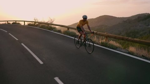 A professional cyclist in a helmet and sports equipment rides on a mountain serpentine at sunset in the direction of the volcano. Steadicam