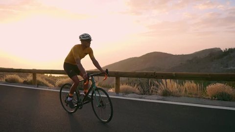 A professional cyclist in a helmet and sports equipment rides on a mountain highway at sunset in slow motion. Steadicam