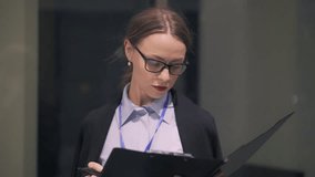 Serious caucasian businesswoman wearing glasses and suit standing in evening street and making notes in her clipboard. Slider slow motion medium shot