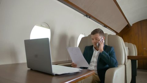 Rich business man talking by mobile phone partner connection aircraft jet travel and work on laptop on luxury expensive table. Middle shot in slow motion.