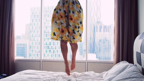 Happy young woman wearing beautiful dress dances jumps on bed at home, slow motion. 3840x2160
