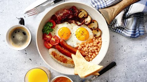 Full English Breakfast served in a pan. Fried eggs, beked beans, tomatoes, champignons, crispy bacon, sausages and toast. Placed on stone background. Top view with copy space.