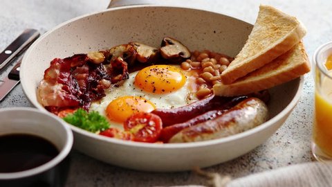 Full English Breakfast served in a pan. Fried eggs, beked beans, tomatoes, champignons, crispy bacon, sausages and toast. Placed on stone background. Top view with copy space. : vidéo de stock