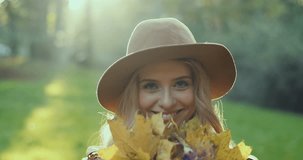 Close up of the smiled charming blonde girl in a hat posing cheerfully in the park and holding yellow autumn leaves and showing them to the camera. Portrait. Outdoor.