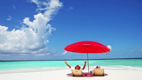 Happy, mid age couple resting on deckchairs under a red parasol at white sand beach in Maldives island