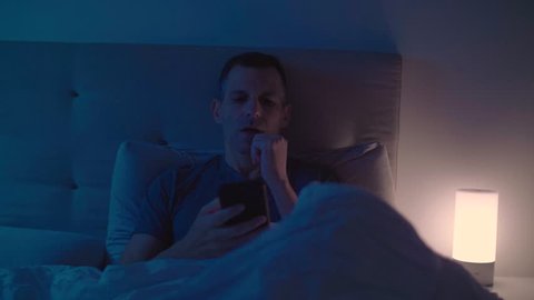 Sleepy guy turns the light off and going to sleep putting down his smartphone beside him