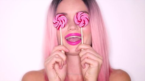 Happy sexy girl playing with lollipop. Beauty Model woman with trendy pink hair style and beautiful makeup holding pink sweet colorful lollipop candy, on white background. Sweets. Slow motion 4K UHD