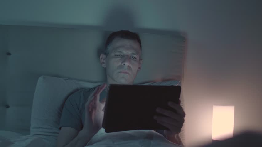 Man Speaking on the phone and looking at the tablet while in bed at night